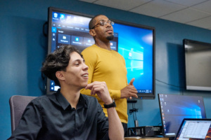 Instructor standing and speaking while a student listens attentively in a classroom with computer monitors.