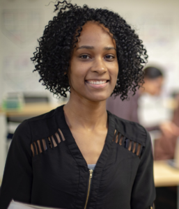 Smiling young woman standing in a classroom with blurred people and whiteboard behind her.