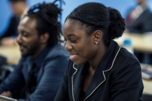 Two adult students in a classroom, a woman smiling and looking down while a classmate works beside her.