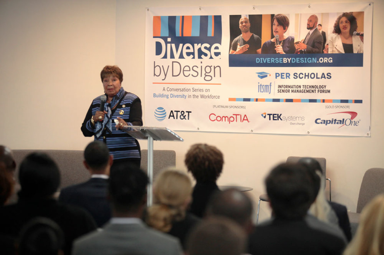 Woman speaking at a podium to a seated audience with a "Diverse by Design" event banner behind her.