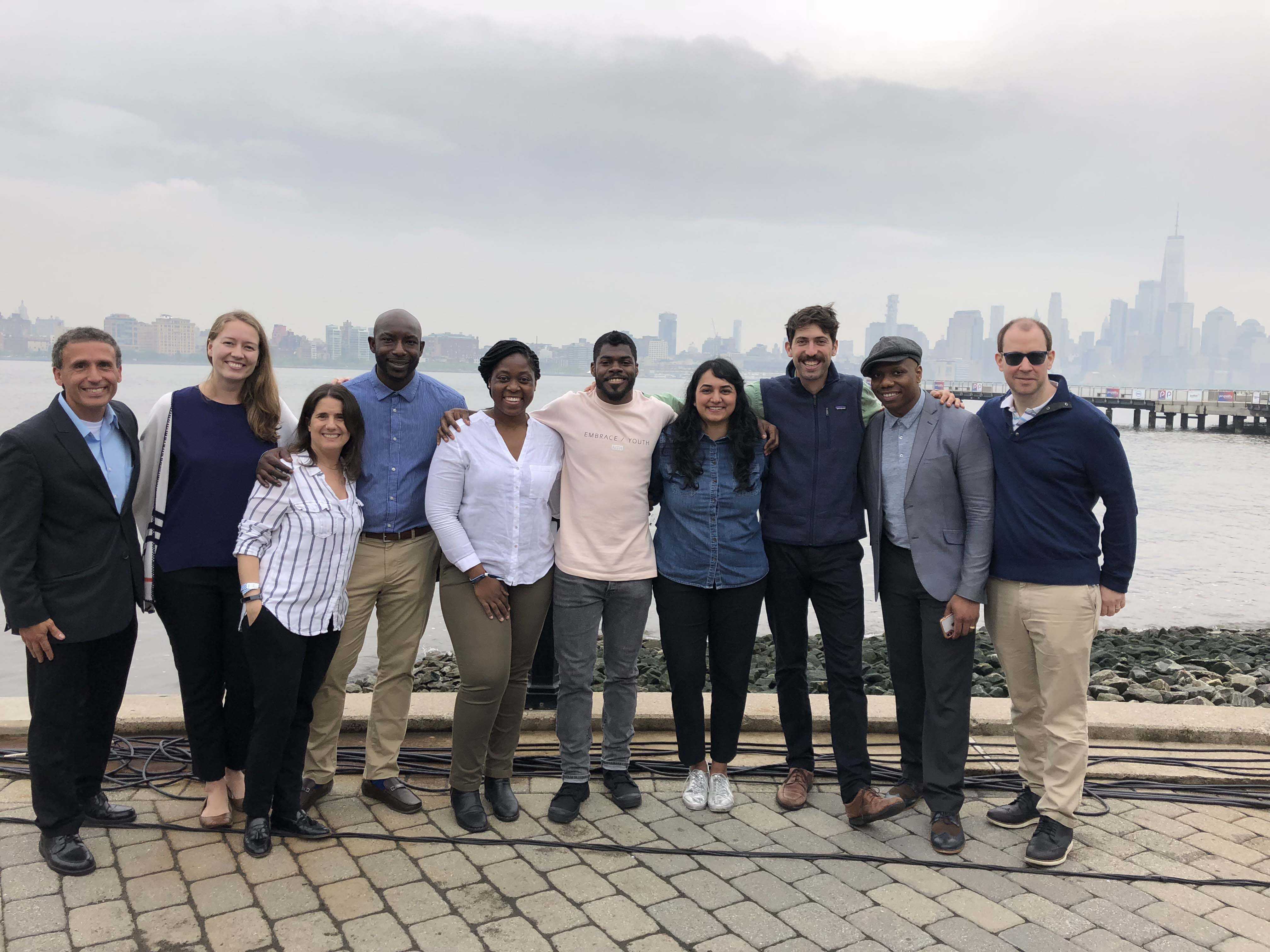 Group of ten adults standing arm in arm by a waterfront, smiling toward the camera with a city skyline behind them.
