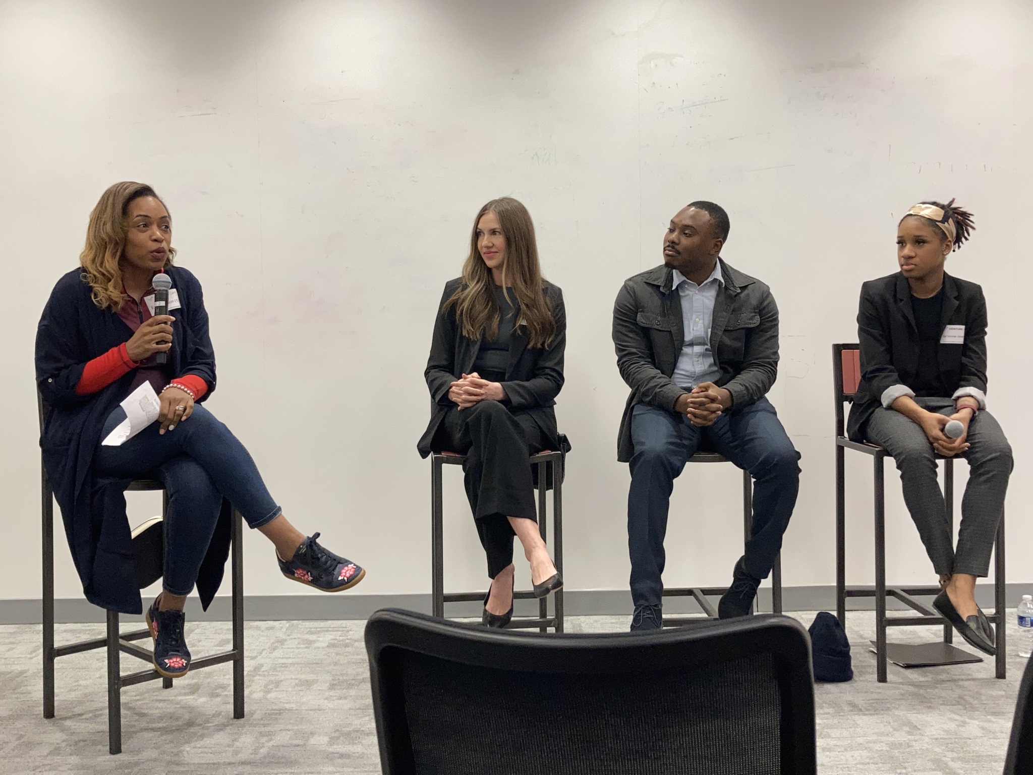 Four panelists seated on stools, one woman speaking into a microphone while the others listen.
