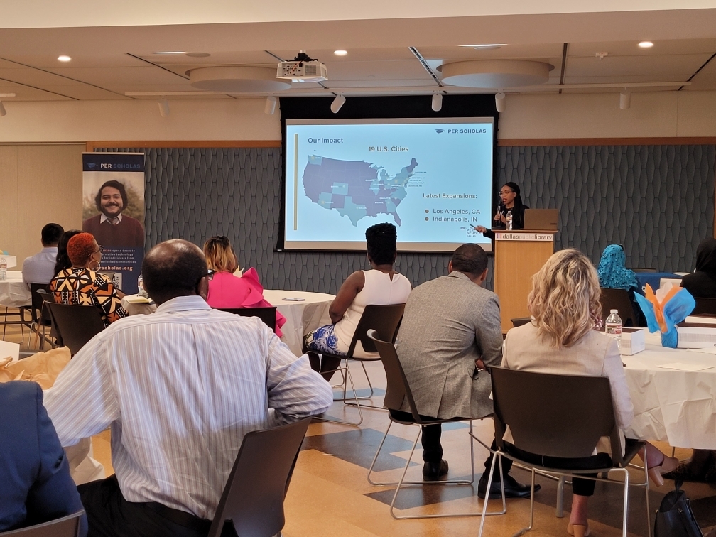 Audience seated at round tables listening to a speaker at a podium presenting a slide about Per Scholas' impact across U.S. cities.