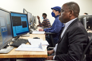 Three adult students focused at desktop computers in a classroom setting.