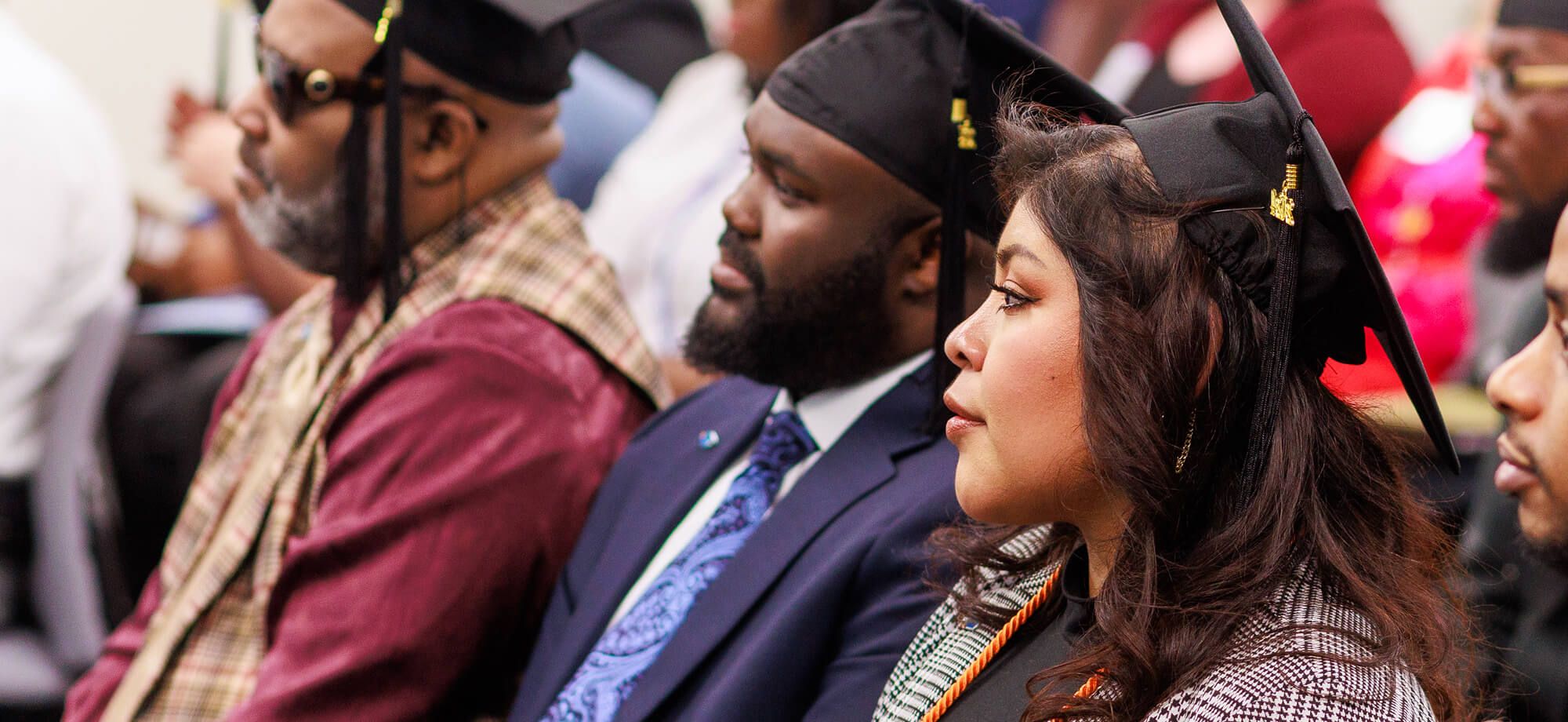 Four adult graduates wearing caps seated in an audience, listening attentively.