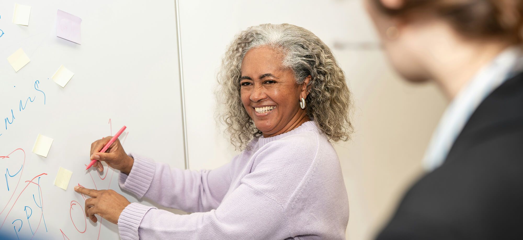 Woman smiling while writing on a whiteboard and pointing to notes, with another person watching.