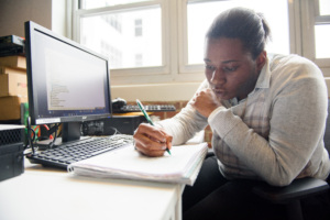 Adult learner focused on writing notes at a desk with a computer displaying code beside them.