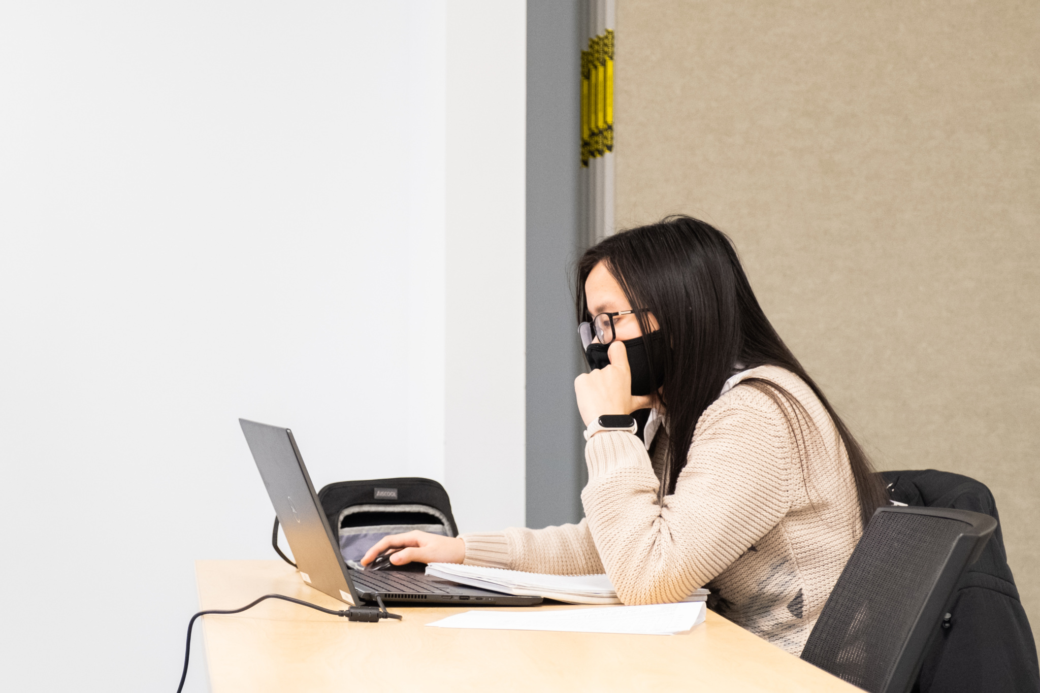 Student wearing a mask works on a laptop at a classroom table, looking at the screen with notes beside them.