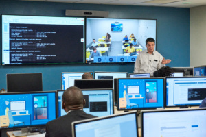 Instructor leading a computer training session with students at monitors and a large screen showing code and a classroom video conference.