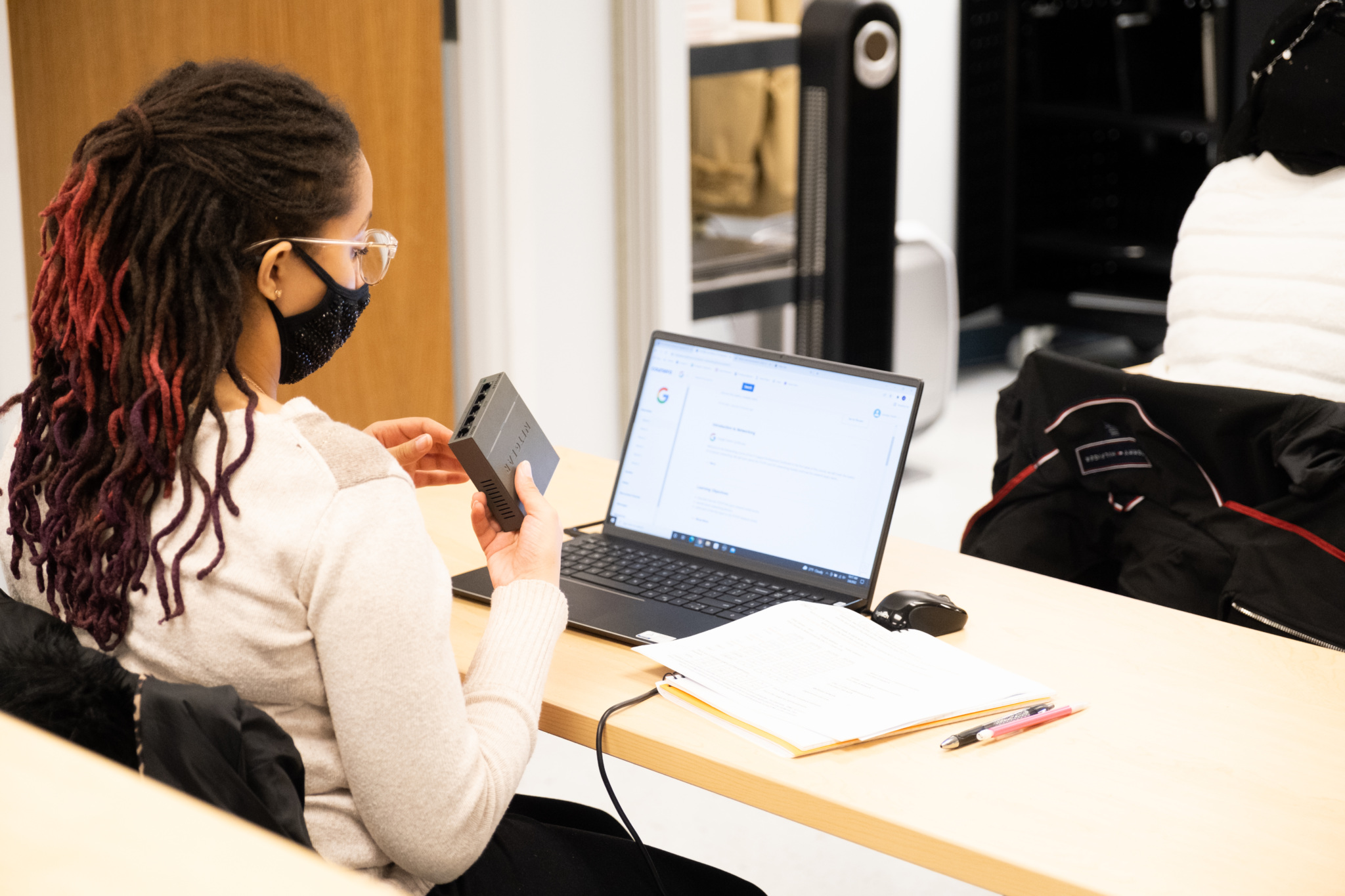 Student wearing a mask works with a small electronic device while looking at a laptop screen in a classroom.
