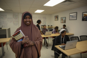 Young adult woman smiles while holding books in a classroom where other students work at laptops.