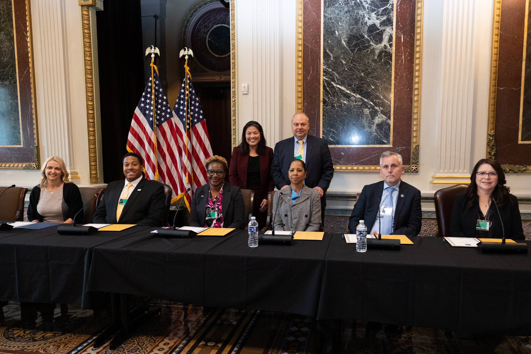 Seven adults seated at a long conference table with two standing behind them and U.S. flags in the background, posing for a photo.