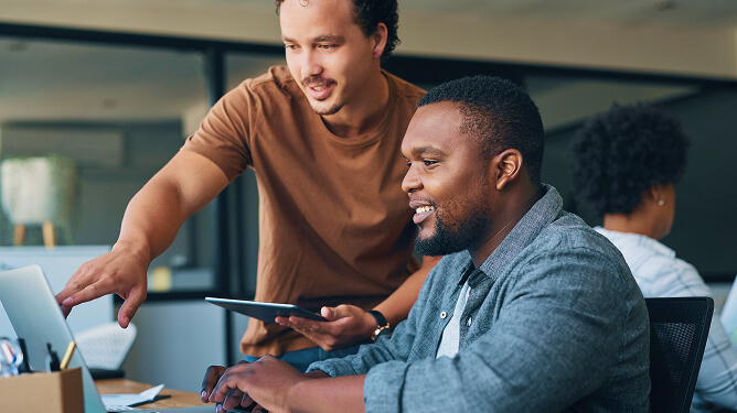 Two male colleagues collaborate at a desk, one pointing at a laptop screen while the other types and smiles.