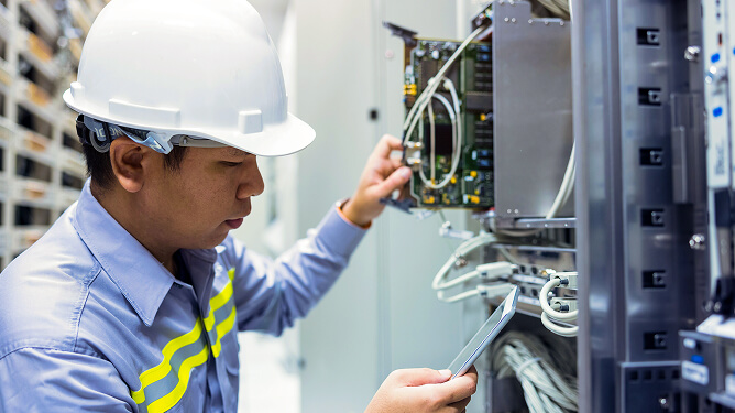 Male technician in a hard hat and safety vest working on network/server hardware while checking a tablet.