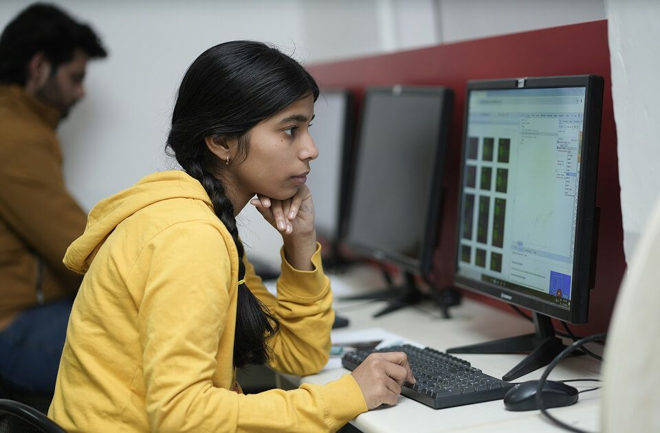 Young woman in a yellow hoodie with braided hair focused on a computer screen while typing, resting her chin on her hand thoughtfully in a computer lab.