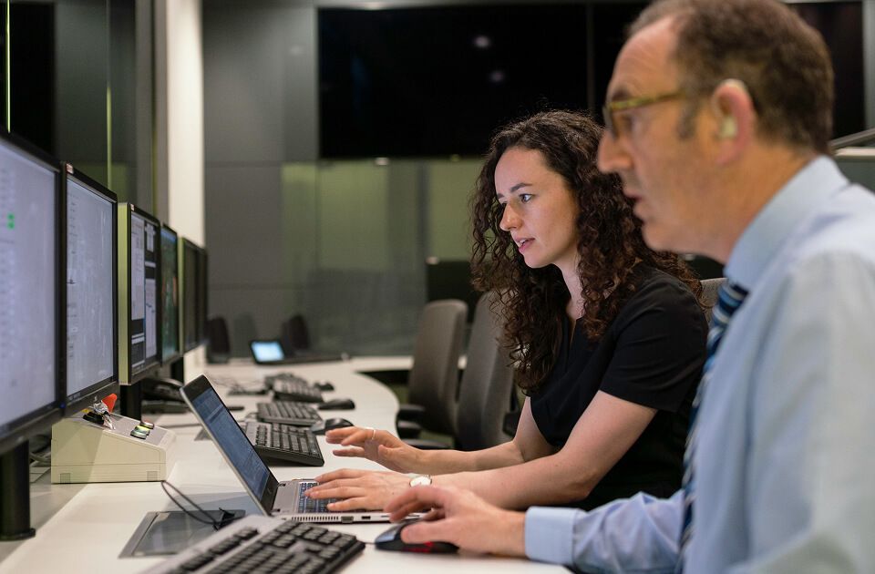 Woman with curly hair typing on a laptop while a man in glasses looks on and speaks, with multiple monitors in a room.
