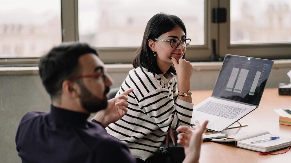 Two coworkers collaborate in a bright office, discussing charts on a laptop; the woman appears thoughtful while the man gestures.