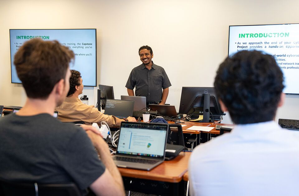 Male teacher at front of classroom instructing group of students working on laptops.