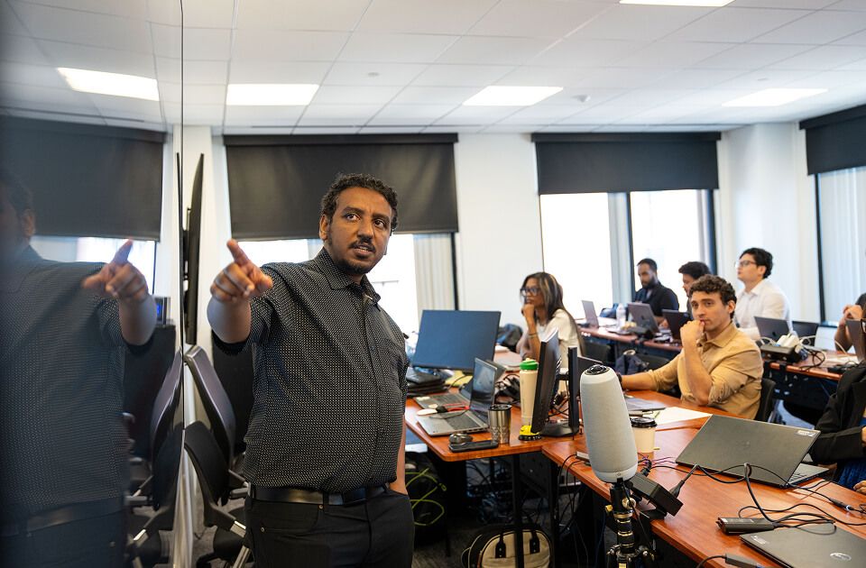 Instructor gesturing while leading a training session as learners at laptops in a conference room listen and take notes.