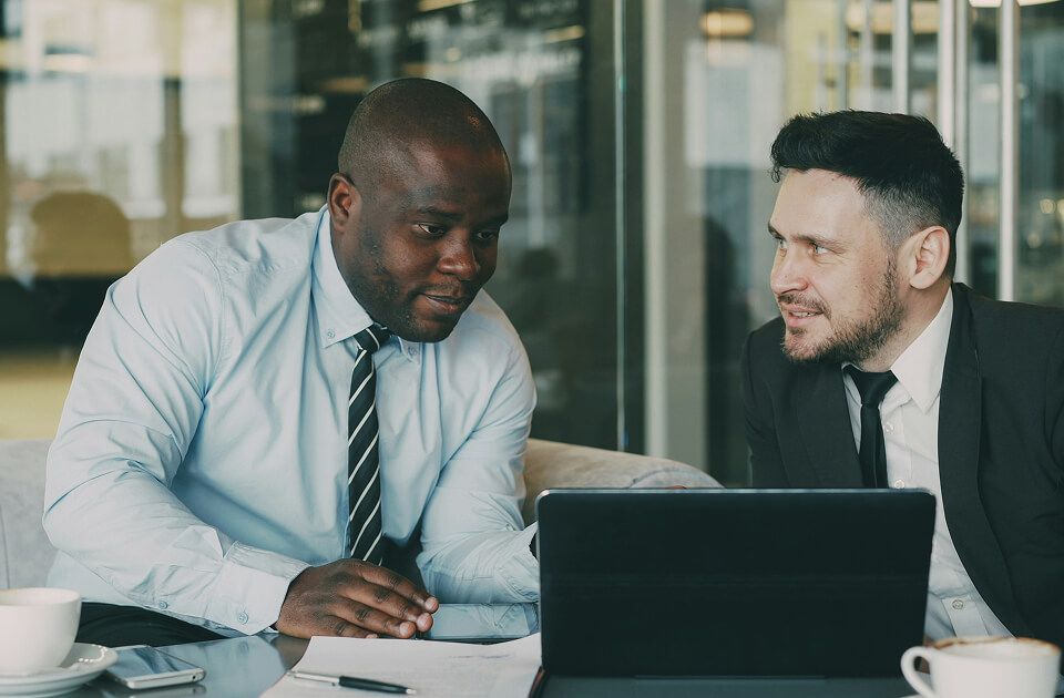Two men in professional attire in conversation over a laptop during a meeting in an office.