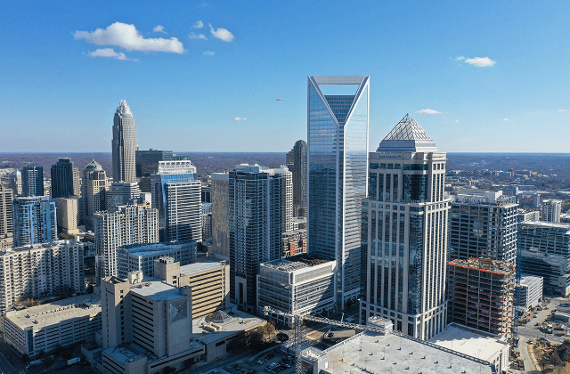 Downtown Charlotte, North Carolina skyline with modern glass skyscrapers under a bright blue sky with scattered clouds.