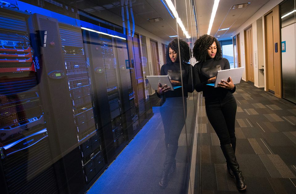 Woman with curly hair holding a tablet standing beside illuminated blue server racks in a data center hallway.