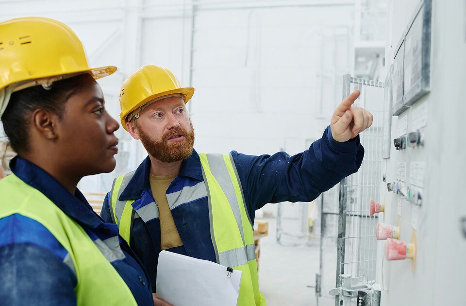 Man in hard hat and safety vest pointing to an electrical panel while explaining to a woman in a hard hat holding a clipboard.