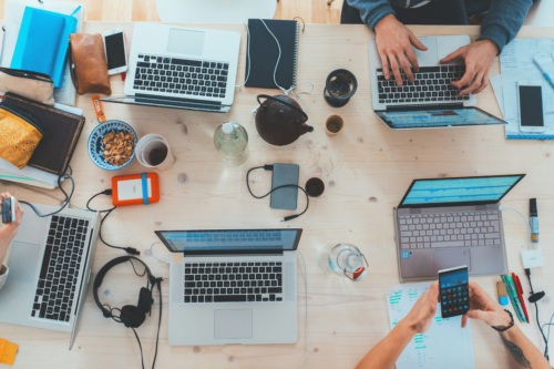A desk seen from above, busy with phones, laptops, and the hands of people working, indicating the importance of mental health awareness in tech.